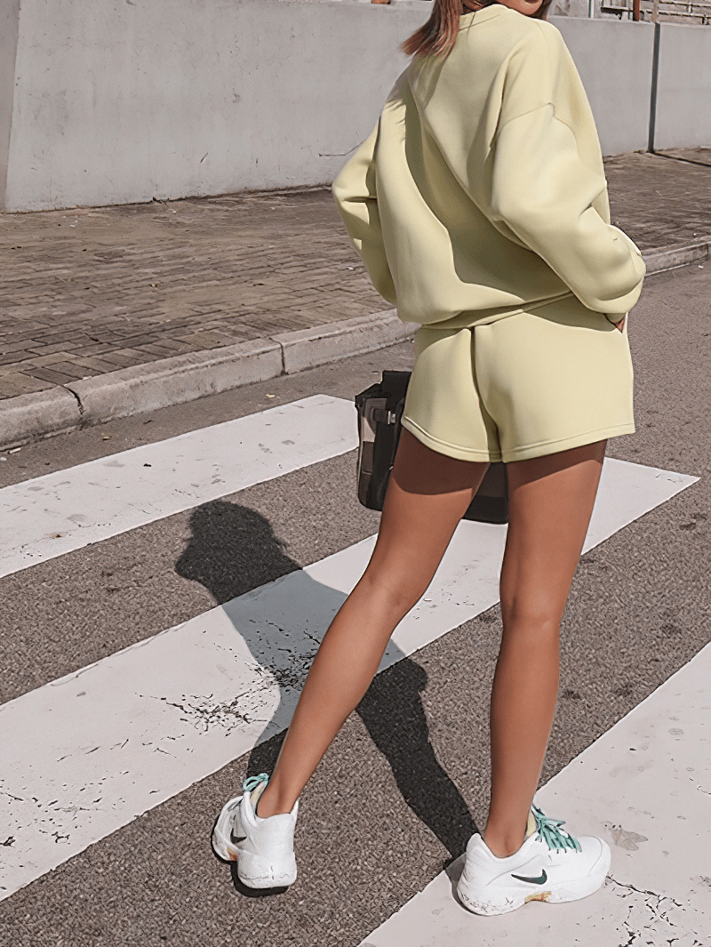 Woman wearing yellow letter embroidery sweatshirt and shorts, standing on a crosswalk, showcasing casual outdoor fashion.